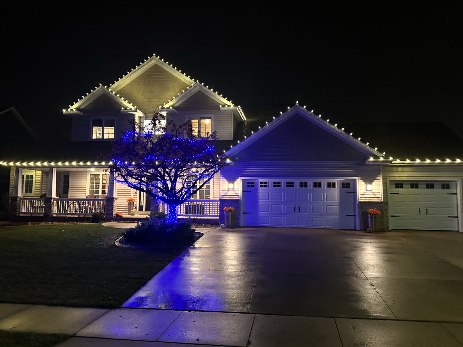 Two-story Rochester home with warm white roofline lights and a blue-lit tree