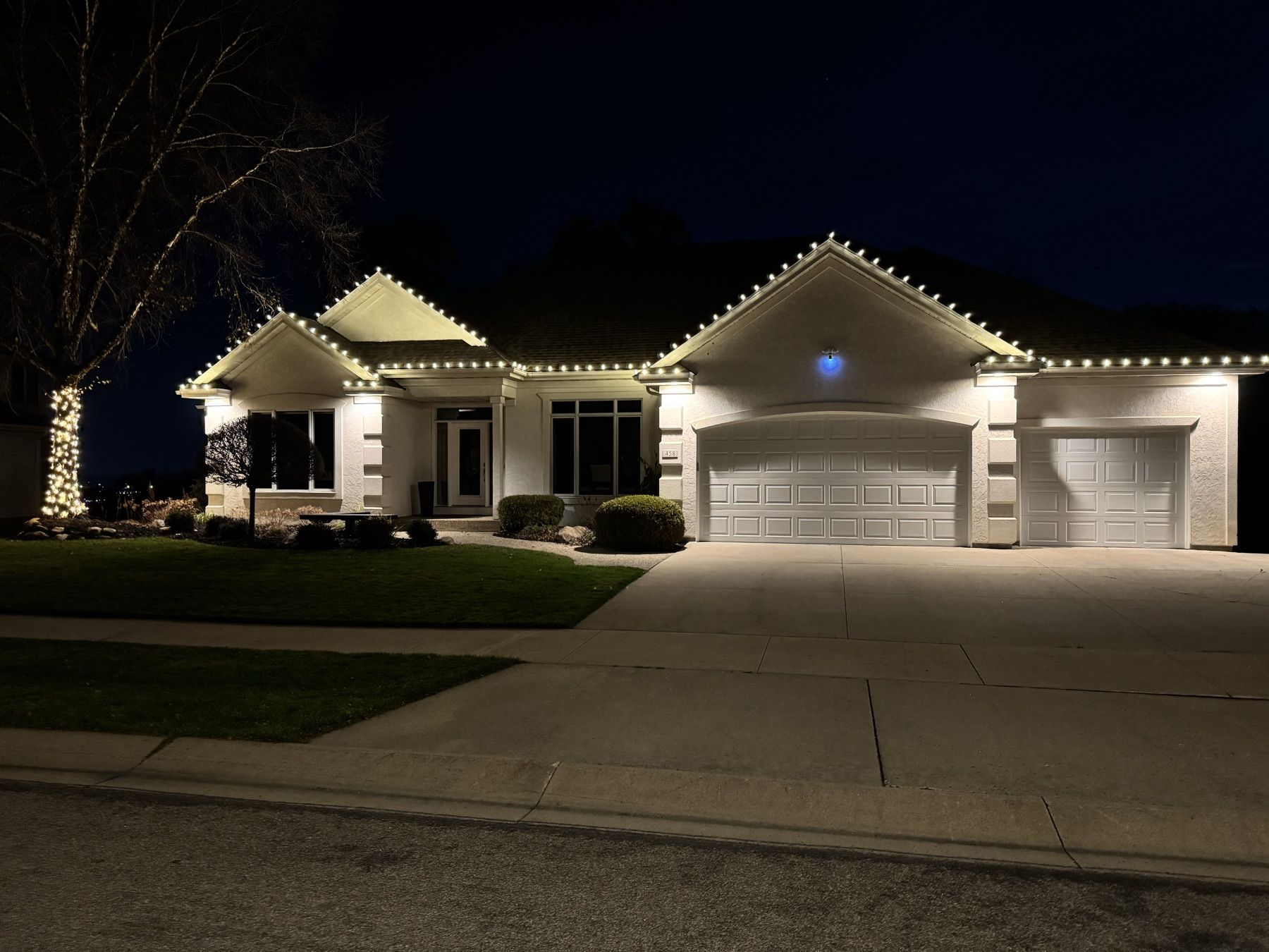 White home with warm white roofline lights and a lit birch tree